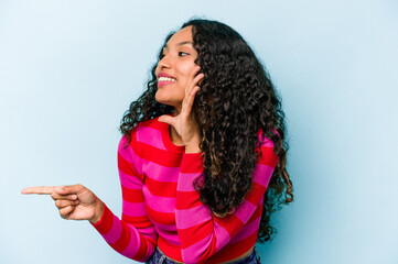 Young hispanic woman isolated on blue background saying a gossip, pointing to side reporting something.