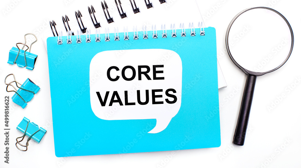 Poster office table with blue notepad, paper clips, magnifying glass and white card with text core values. 