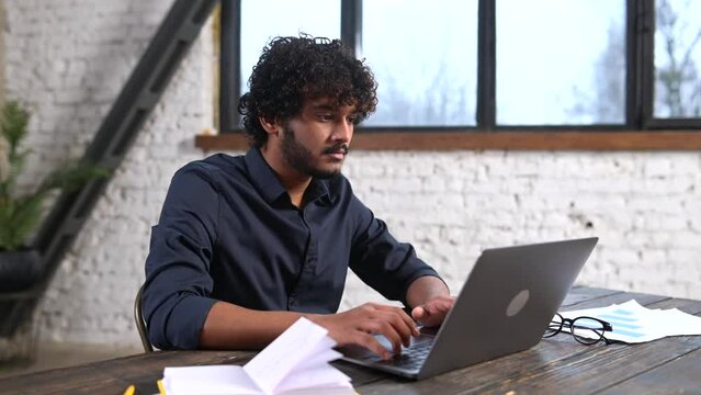 Confident concentrated Indian freelancer guy working on laptop at home office, sitting at desk and using computer. Multiracial eastern man typing on keyboard, looking at screen, enjoying remote work