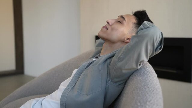 Asian guy leans on armchair back nodding head while listening to dynamic music. Young man relaxes at home on blurred background close side close