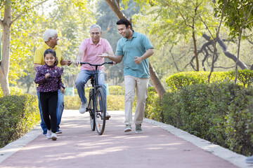 Senior man riding bicycle with granddaughter running while other old man and son encouraging at park