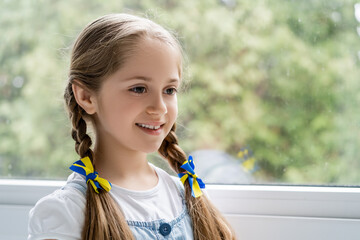 patriotic girl with blue and yellow ribbons on braids smiling near blurred window.