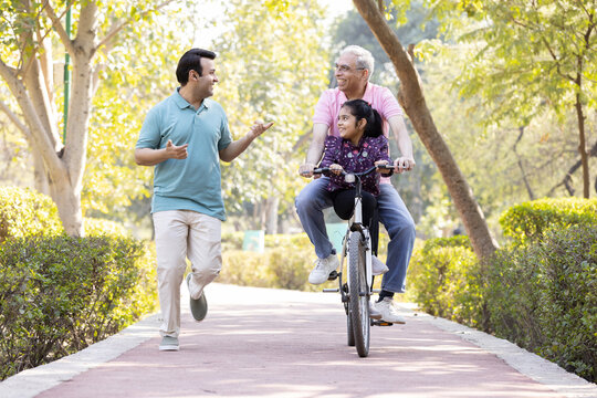 Cheerful senior man riding bicycle with granddaughter while son running at park