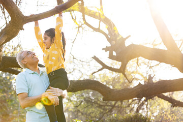 Cheerful senior man helping granddaughter hanging from tree branch at park - obrazy, fototapety, plakaty