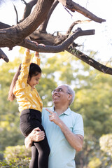 Cheerful senior man helping granddaughter hanging from tree branch at park