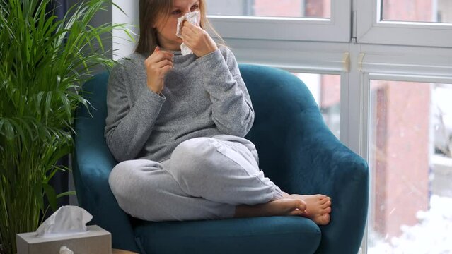 Unhealthy woman sits in a chair and coughs because she has a cold, flu, coronavirus. It is snowing outside