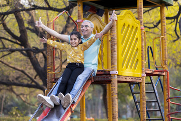 Senior man with granddaughter having fun while sliding in playground at park