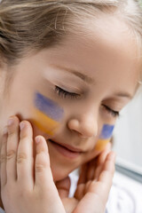Obraz na płótnie Canvas close up portrait of girl with closed eyes touching face with painted ukrainian flags.