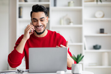 Smiling attractive millennial arab male with beard in red clothes speaks by phone and looks at...