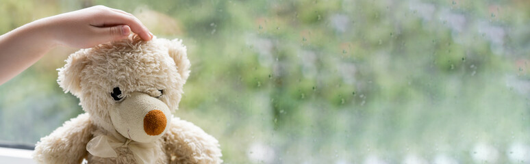 partial view of hand of child near teddy bear and blurred window with raindrops, banner.