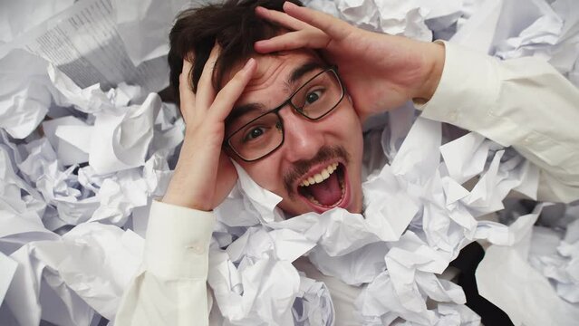 Male office worker grabs head and screams while in pile of crumpled papers. Man screams being among lot of crumpled documents