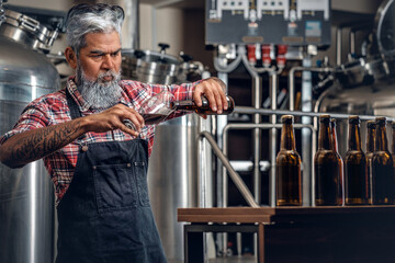 Shot of bearded senior brewer pouring his product to glass around table with beer and barrels.