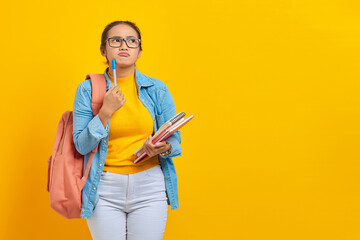 Portrait of serious young Asian woman student in casual clothes with backpack holding book and pen, thinking about question isolated on yellow background. Education in college university concept