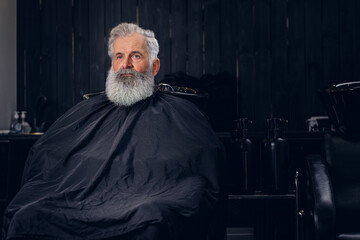 Shot of fashionable old hipster with long beard and peignoir in dark barber shop.