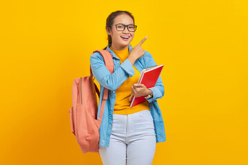 Portrait of smiling young Asian woman student in casual clothes with backpack holding book and pointing aside with finger isolated on yellow background. Education in college university concept