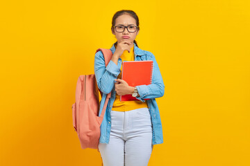 Portrait of pensive young Asian woman student in casual clothes with backpack holding book and thinking about question isolated on yellow background. Education in college university concept