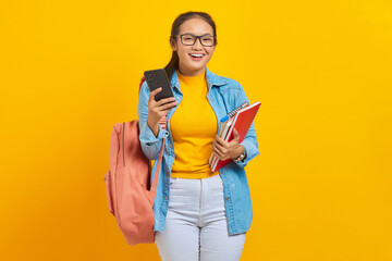Portrait of cheerful young Asian woman student in denim clothes, glasses with backpack holding mobile phone and books isolated on yellow background. Education in high school university college concept