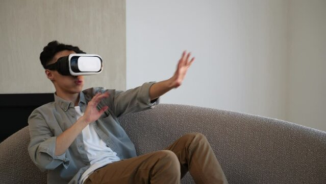 Asian man in goggles enjoys virtual reality getting scared of terrifying images. Young guy in casual clothes sits on sofa at home moving hands