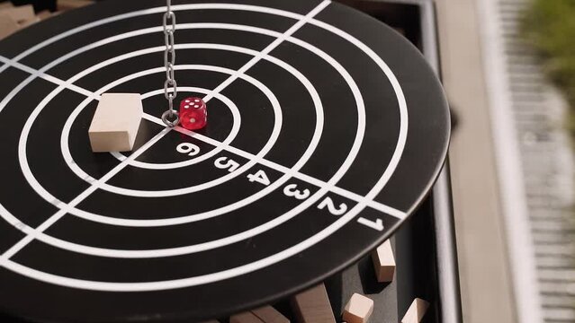 Round black disc of the balance board game hangs on chain and sways over the playing field. On black circle with markings lies a wooden block and a red dice with numbers.