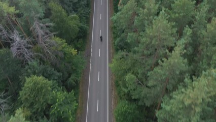 Aerial shot to biker riding on motorbike through forest road. Motorcyclist racing his motorcycle on country road at green wood. Man drive bike during trip. Concept of freedom and adventure. Top view