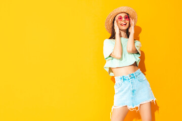 Portrait of young beautiful smiling female in trendy summer jeans skirt. carefree woman posing near yellow wall in studio. Positive model having fun indoors in sunglasses. Cheerful and happy. In hat