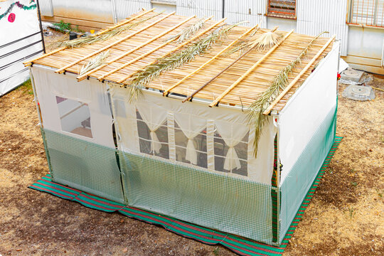 White Sukkah outside on the street with a garden and trees near the building, a traditional building for the Jewish holiday of Sukkot. Jewish tradition. Traditional hut for the celebration of Sukkot. 