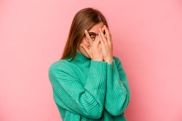 Young caucasian woman isolated on pink background blink through fingers frightened and nervous.