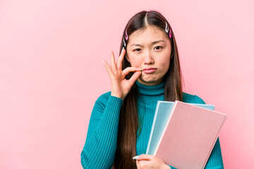 Young asian student woman holding books isolated on pink background with fingers on lips keeping a secret.