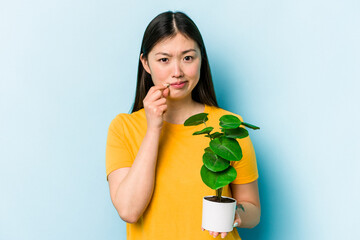 Young asian woman holding a plant isolated on blue background with fingers on lips keeping a secret.