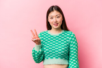 Young Chinese woman isolated on pink background showing victory sign and smiling broadly.