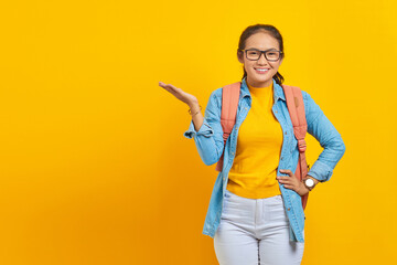 Smiling young Asian woman student in denim outfit with backpack pointing to copy space with palms,...