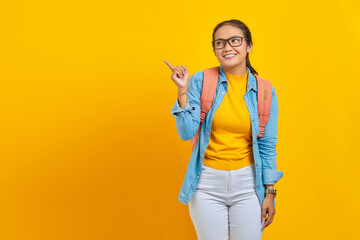 Smiling young Asian woman student in denim outfit with backpack pointing finger in copy space, showing advertising products isolated on yellow background.  Education in university college concept