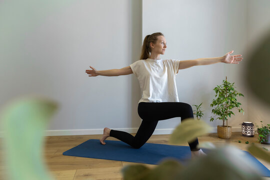 young slim woman is practicing yoga, twisting chest, beneficial for back, standing on one knee, looking forward, wearing beige oversize t-shirt and black leggings, blurred plants on the foreground