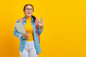 Portrait of cheerful young Asian woman student in casual clothes with backpack holding laptop and showing ok gesture with finger isolated on yellow background. Education in university college concept