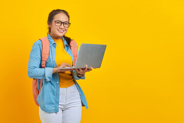 Portrait of cheerful young Asian woman student in casual clothes with backpack using laptop and looking at camera isolated on yellow background. Education in university college concept