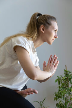 A young Caucasian female with blonde hair in a tail, holds her hands in namaste or prayer, religious greeting, looking calmly in front of her, practicing yoga balanced pose
