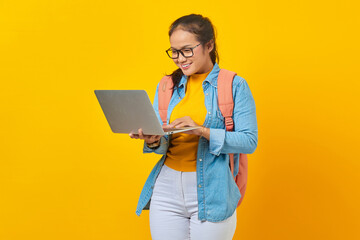 Fototapeta na wymiar Portrait of smiling young Asian woman student in casual clothes with backpack looking at incoming email on laptop isolated on yellow background. Education in university college concept
