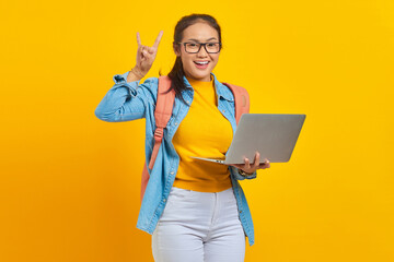Portrait of cheerful young Asian woman student in casual clothes with backpack using laptop and doing rock symbol with hands up isolated on yellow background. Education in university college concept