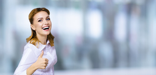 Excited happy smiling businesswoman, showing thumb finger up or like hand sign gesture. Success in business studio concept. Blurred modern office background.