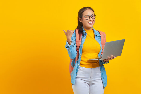 Portrait Of Smiling Young Asian Woman Student In Casual Wear With Backpack Using Laptop And Pointing At Copy Space With Finger Isolated On Yellow Background. Education In College University Concept