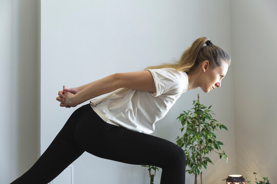 young slim woman is stretching her arms, opening chest, holding arms behind, leaning forward to her leg, profile portrait of practice at home