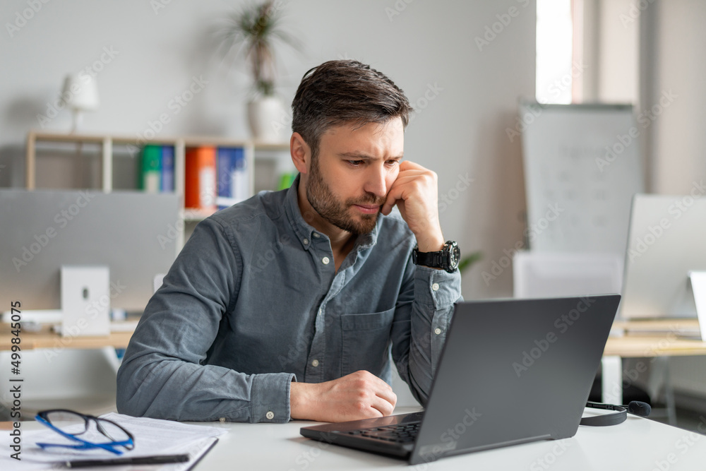 Poster serious middle aged businessman using laptop computer and reading reports, sitting at workplace in o