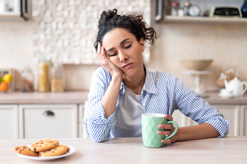 Sleepy young woman sitting at dining table in kitchen with closed eyes, holding cup while having...