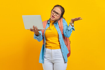 Portrait of angry young Asian woman student in casual clothes with backpack talking on smartphone while holding laptop,saying no with palms isolated on yellow background