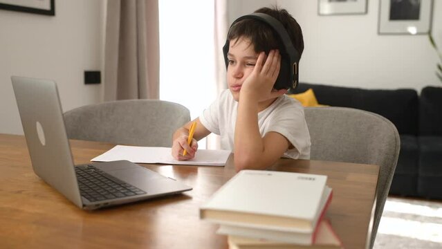 Tired schoolboy doing homework and listening music in the headphones, studying on the distance from home, using trendy laptop, preteen boy writing tasks in the notebook. Online education concept