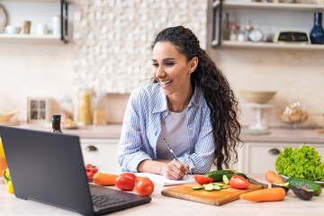 Writing down new recipe. Happy latin woman taking notes in copybook while watching cooking webinar on laptop