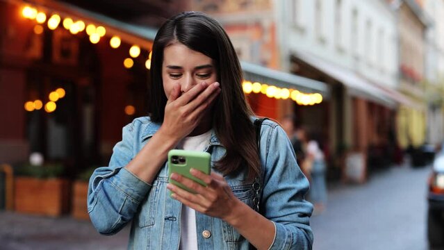 Happy girl reading good news in message outdoor. Portrait amazed woman receiving good news at street. Exited lady celebrating victory with smartphone at street