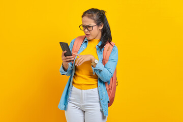 Portrait of  surprised young Asian woman student in casual clothes with backpack typing text message or scrolling feed on social network using smartphone isolated on yellow background