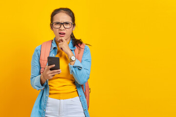 Portrait of pensive young Asian woman student in casual clothes with backpack using mobile phone and holding chin while thinking something isolated on yellow background