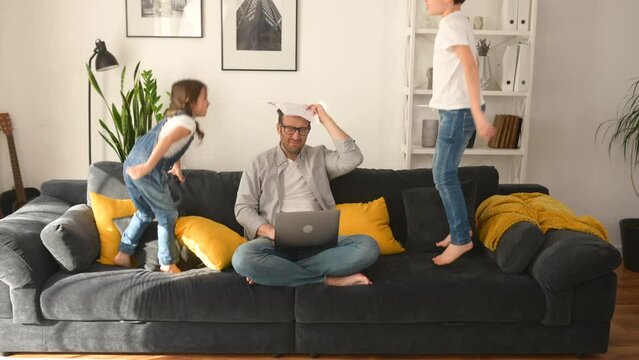 Crazy irritated father screaming with eyes closed, while his two fidget kids are shouting, jumping on the sofa around. Stressed busy single parent is babysitting and working on the distance